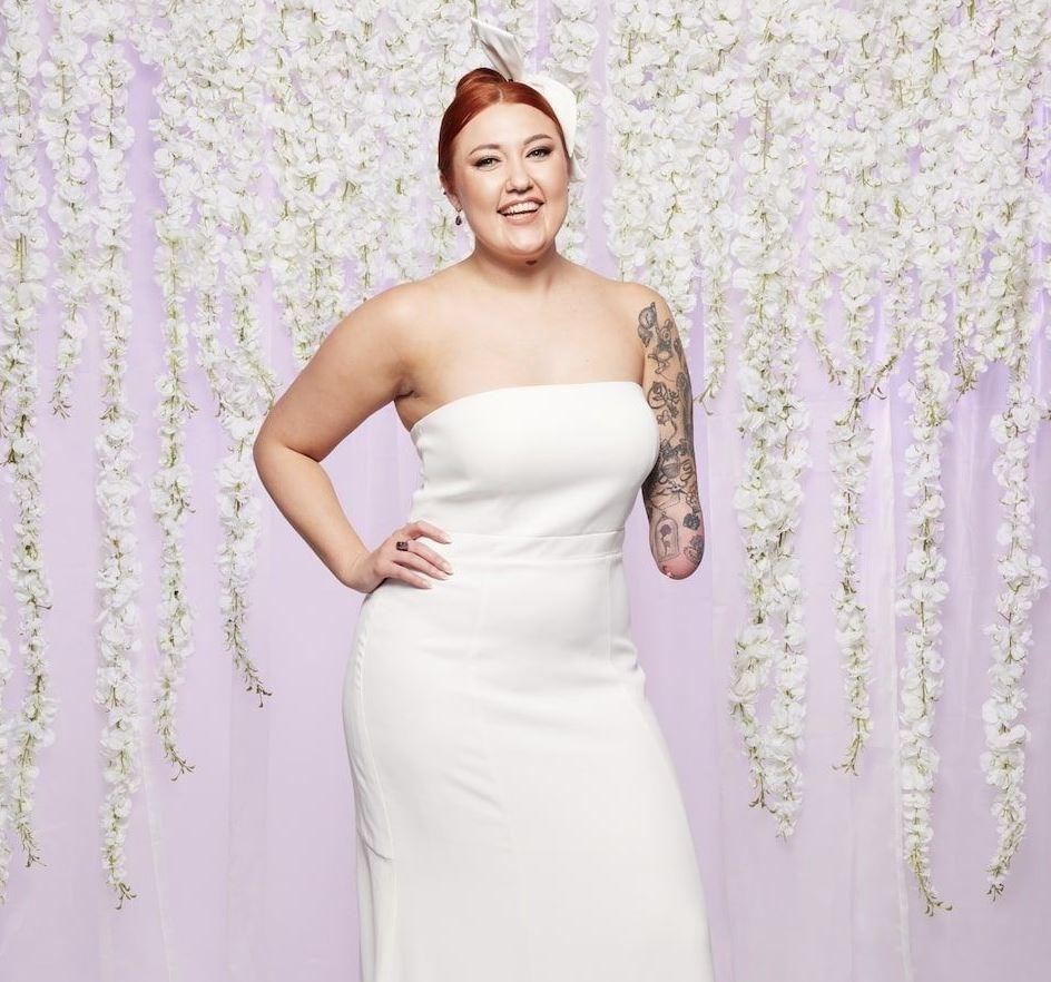 mafs uk bride jay wears a strapless white wedding dress and smiles with her hair up in front of lilac backkdrop