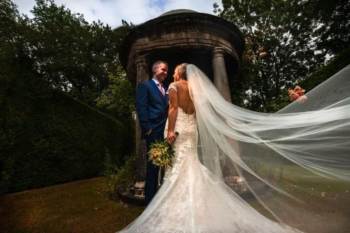 a view of the back of a bride's open back wedding dress and floating veil as she and her groom stand at the altar outdoors