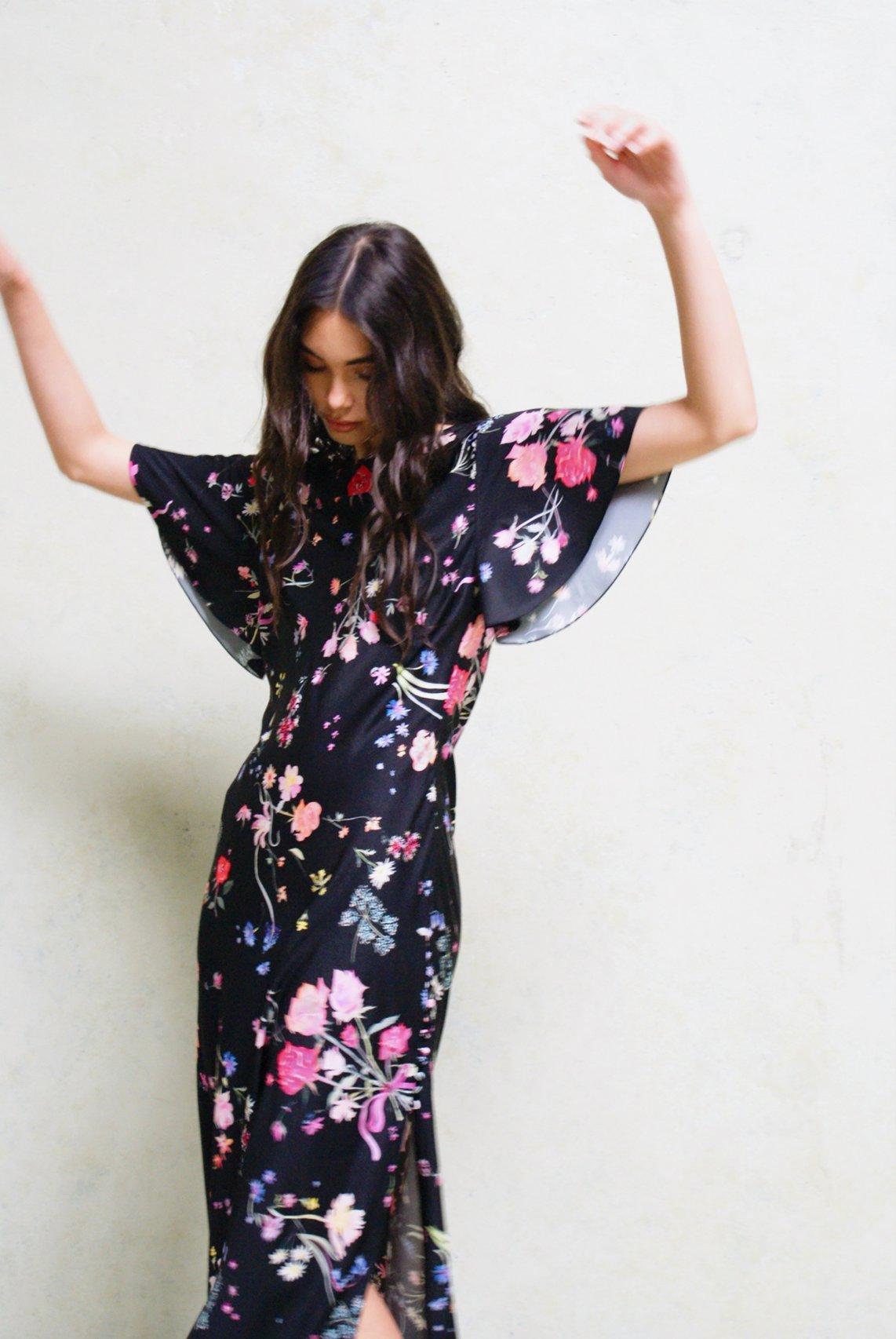 A wedding guest standing with her hands in the air wearing a black maxi dress covered with a pretty floral print. The dress has short fluted sleeves.