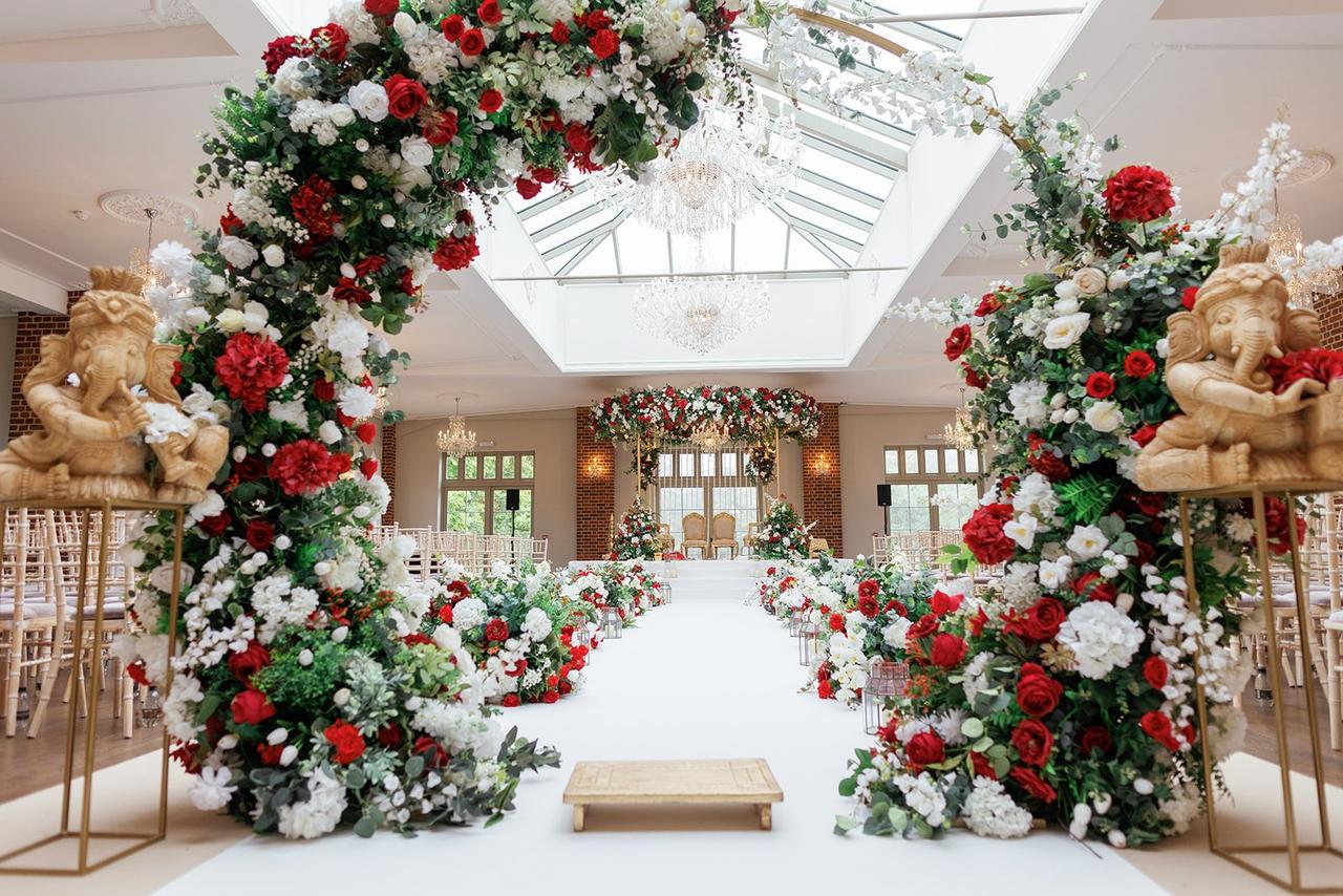 Interior of a country house wedding venue set up for an Asian wedding ceremony with colourful floral-lined aisles and grand chairs.