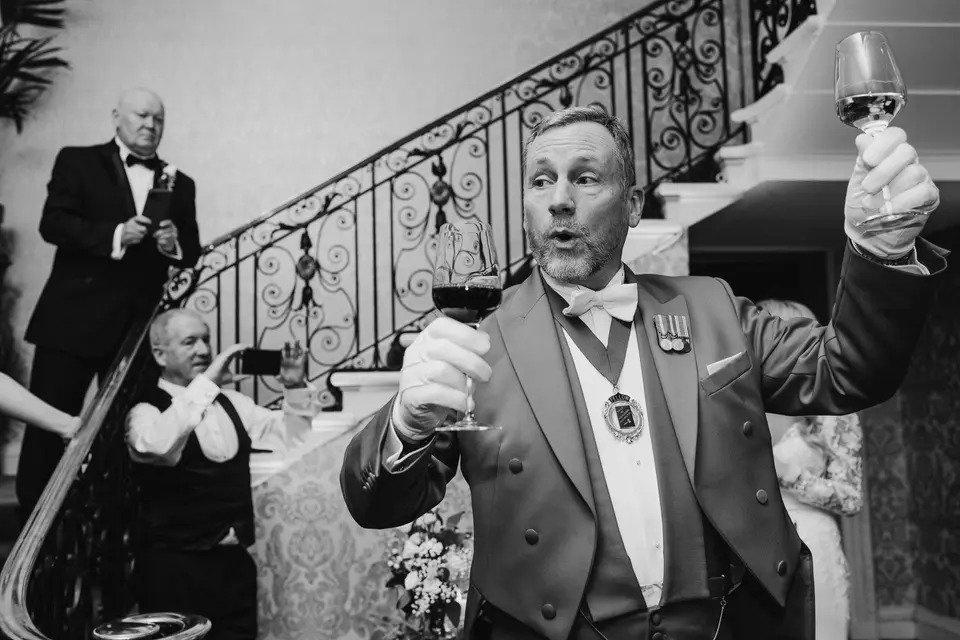 Black and white photo of a wedding toastmaster holding up a glass of red wine and a glass of white wine