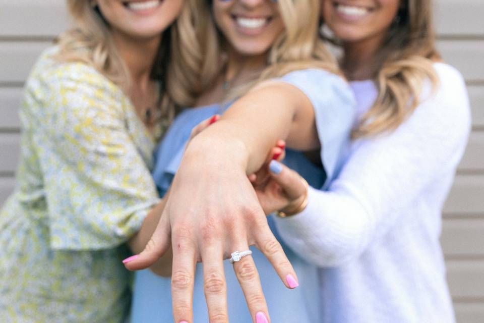A bride to be holding out her ring finger revealing a diamond engagement ring as a friend stands either side of her hugging her