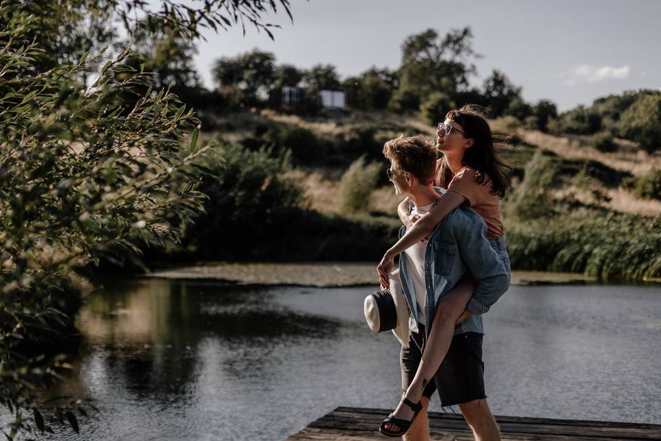 A man is piggyback carrying a woman as they walk past a lake and look at the scenery