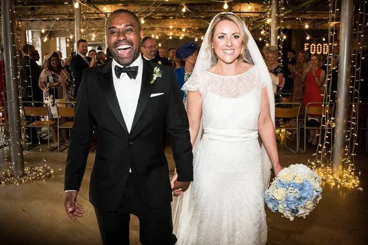 An ecstatic bride and groom leaving their wedding ceremony room hand in hand beaming. The groom wears a black tuxedo and a bow tie and the bride wears a simple wedding dress and veil and carries a bouquet of blue and white flowers. Their guests are visible in the fairy light strewn, rustic ceremony space behind them, all applauding 