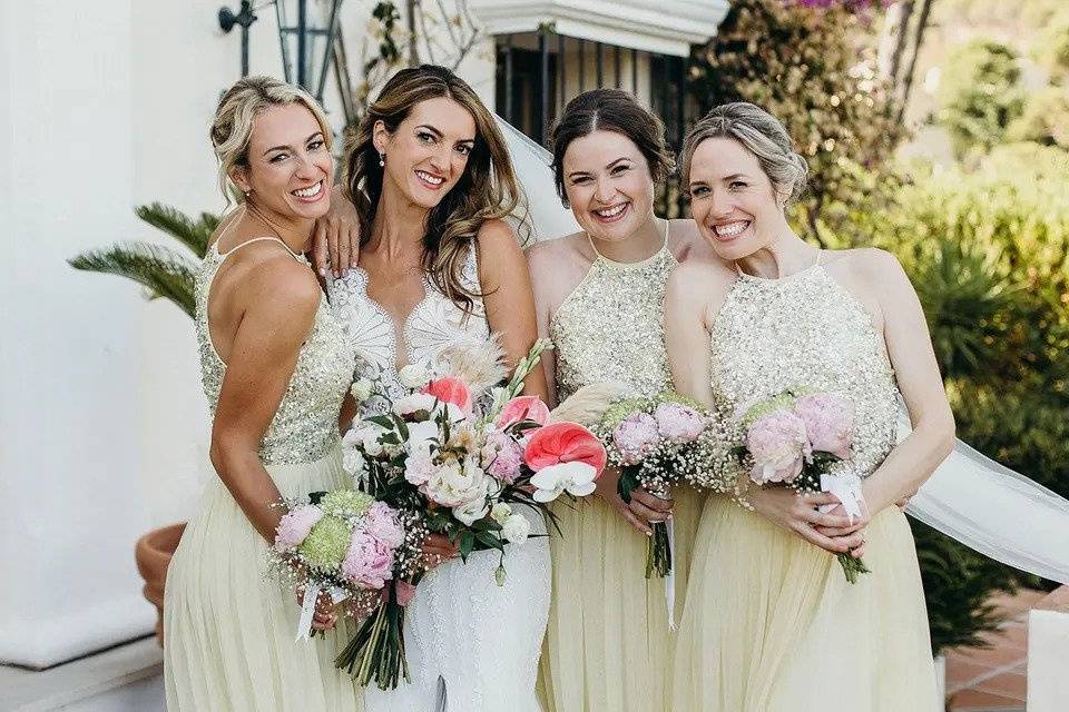 A bride holding a bouquet poses with her bridesmaids, also holding bouquets, outdoors with a white wall and plants in the background