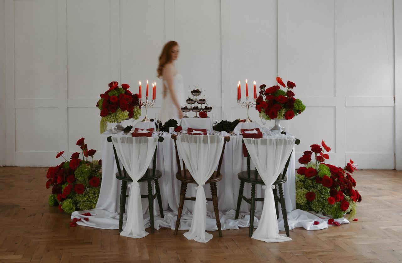 bride walking past a long white wedding table covered in white tulle and red roses