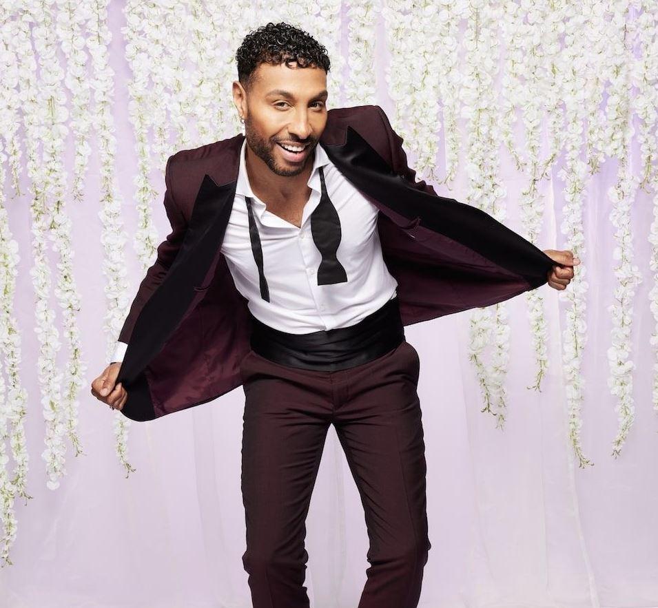 mafs uk groom nathanial wears a burgundy suit and has his bow tie undone in front of a lilac backdrop with white hanging flowers