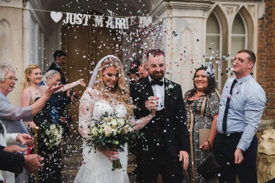 Bride and groom exit wedding venue holding hands while their guests throw confetti over them