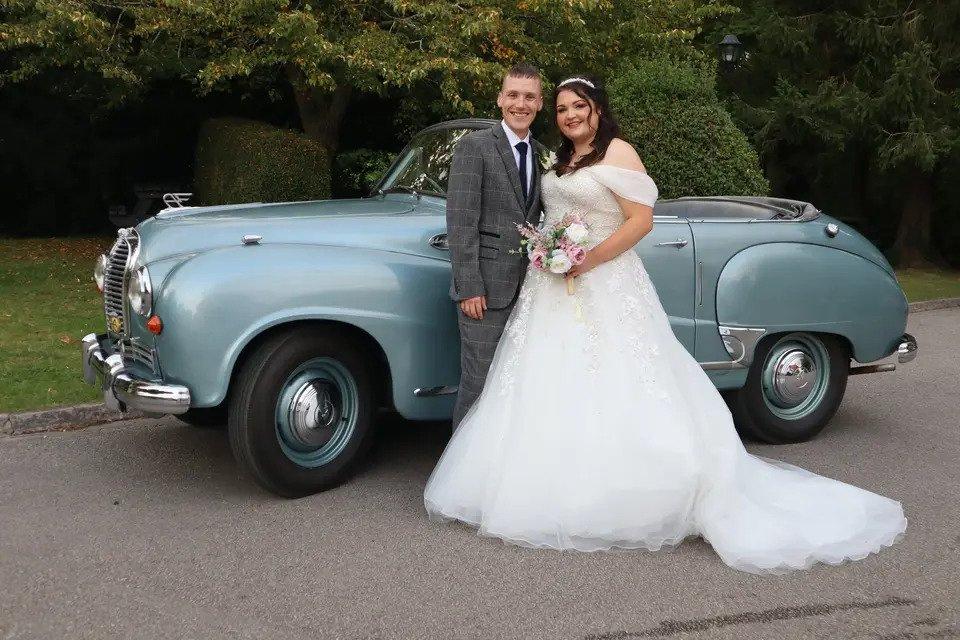 Bride and groom smile at the camera in front of a classic car