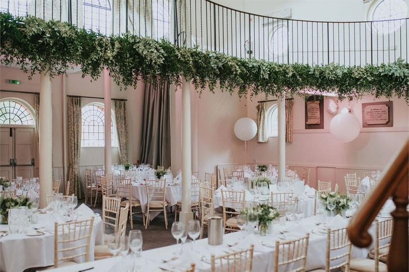 Chapel room decorated for wedding meal with round white tables, banquet table, Chiavari chairs, foliage decor, floral centrepieces, lanterns and a higher level with black railings