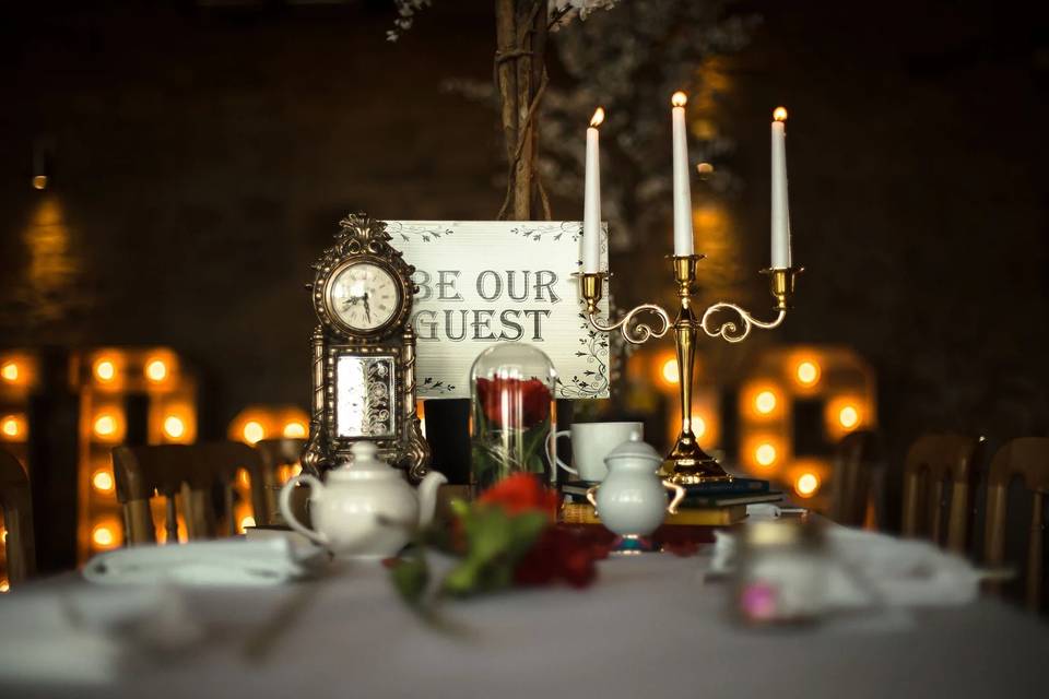 Wedding table with a clock, red rose, gold candelabra and be our guest sign 