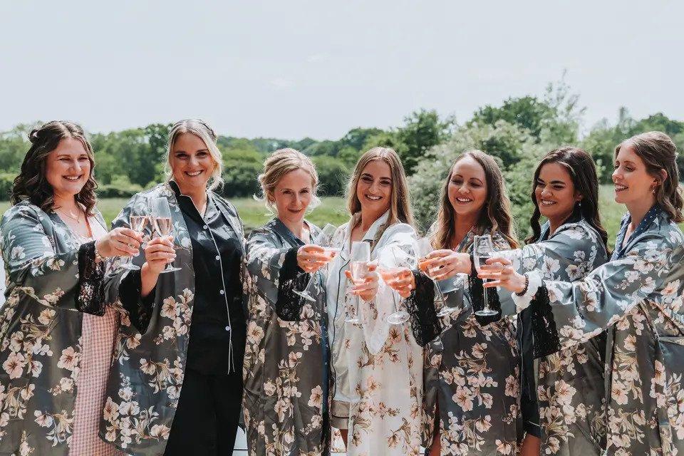 Bride and six bridesmaids posing outside in their PJs and matching floral bridal dressing gowns, holding out their glasses of champagne as though to cheers each other