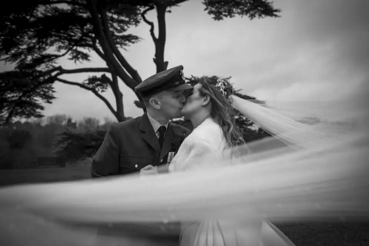a couple kissing in a black and white picture in victorian-style wedding attire as the bride's veil swooshes in front of the camera