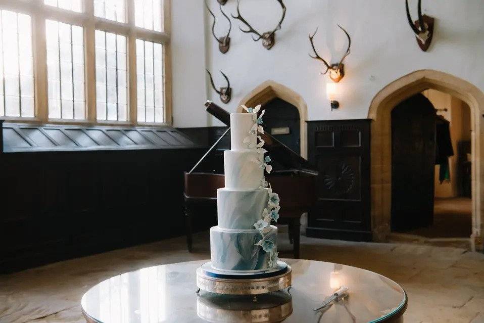 Four tier ombre marble cake starting as white and descending to a deep blue with white and blue sugar flowers. The cake is on top of a table in a room with a grand piano and mounted stags heads