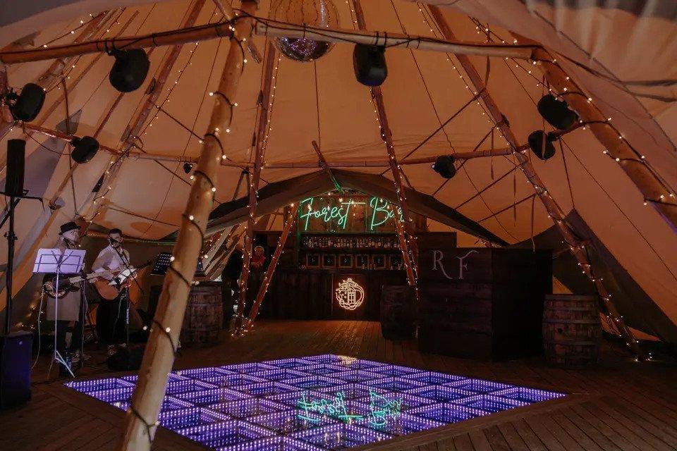 interior of a tipi wedding venue with purple light up dance floor, fairy lights, wooden beams and neon sign reading 