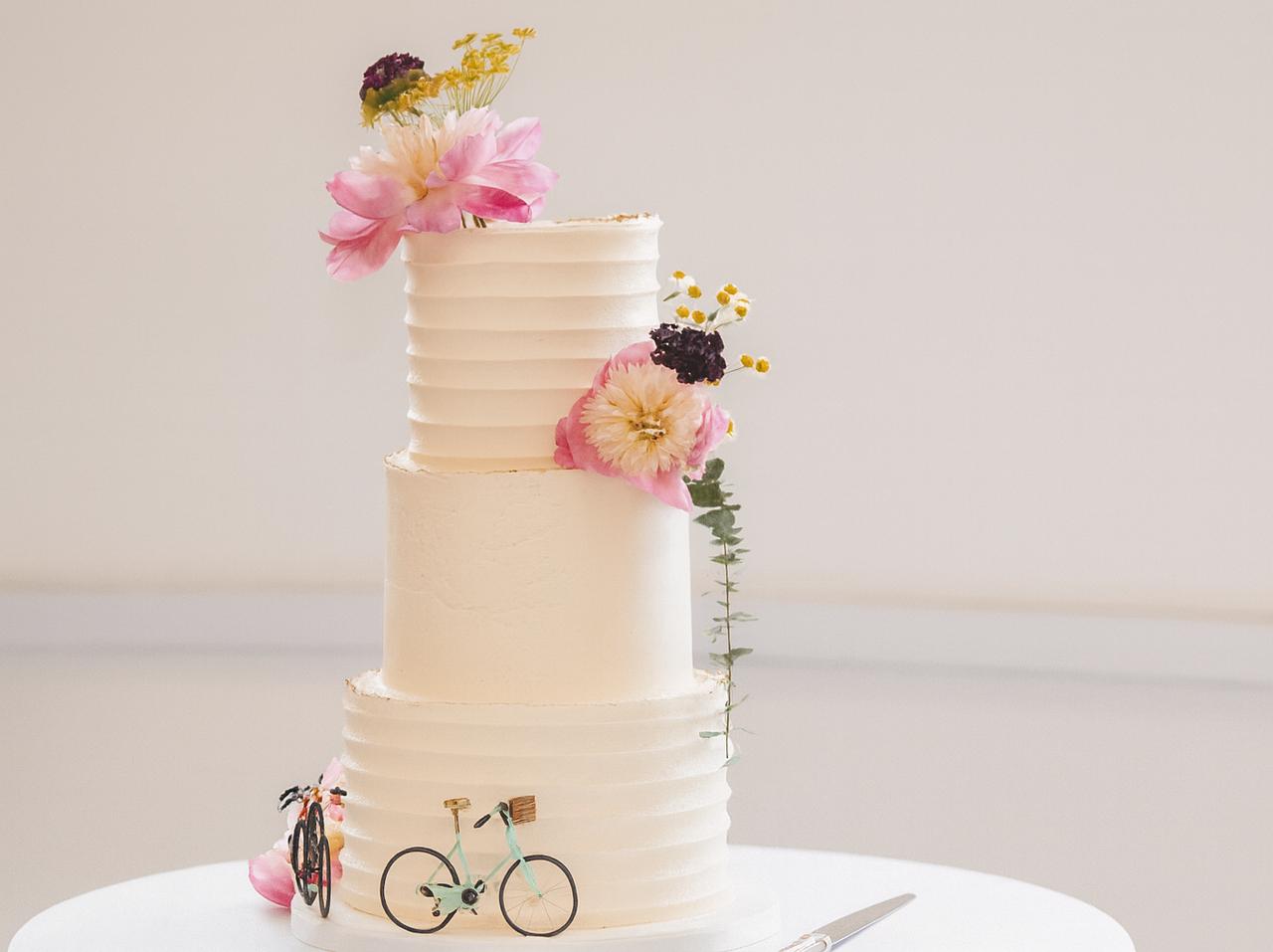 A simple three tier white wedding cake decorated with a few pink flowers and bicycle decorations.