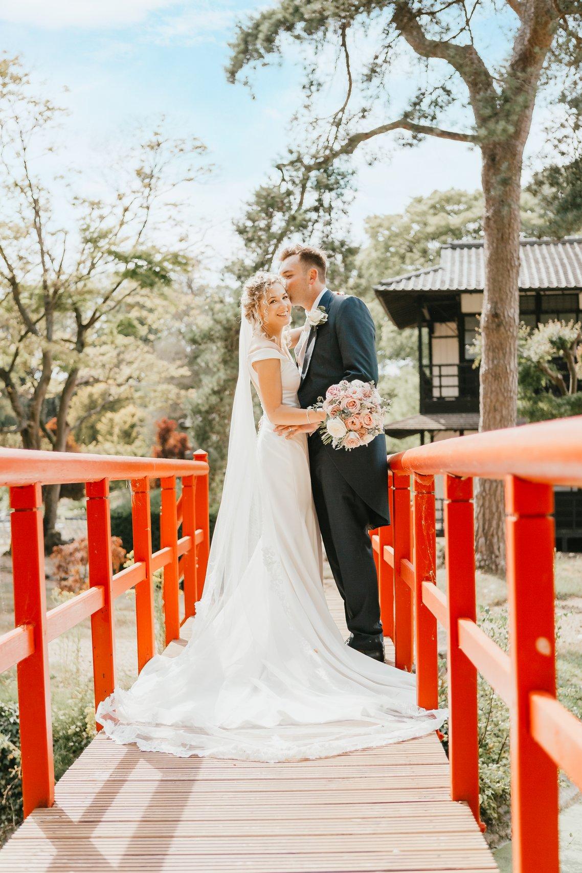 The groom kisses the brides cheek on a red bridge