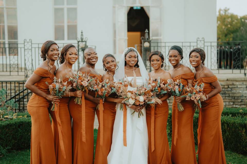 a bride in a white dress and veil with her seven bridesmaids in orange dresses all holding orange flower bouquets