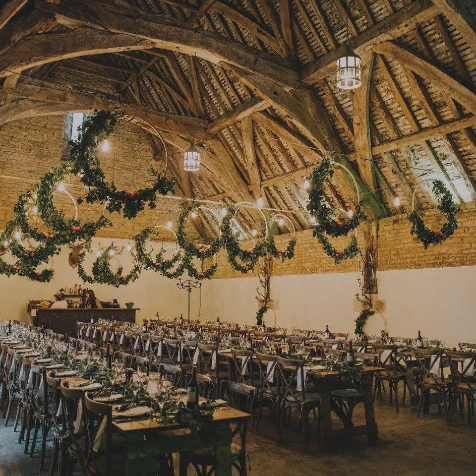 Barn interior laid for wedding breakfast with long tables, wood chairs and exposed beams and foliage hoops
