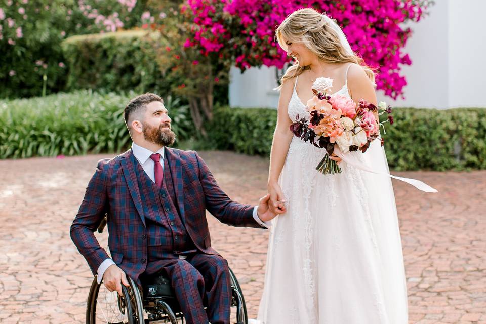 Bride stands and holds her husband's hand as he sits in a wheelchair outside a wedding venue in South Africa