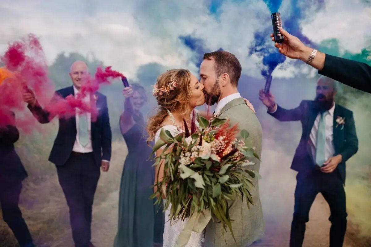 a bride kissing her groom as members of the wedding party wave pink purple and blue smoke around
