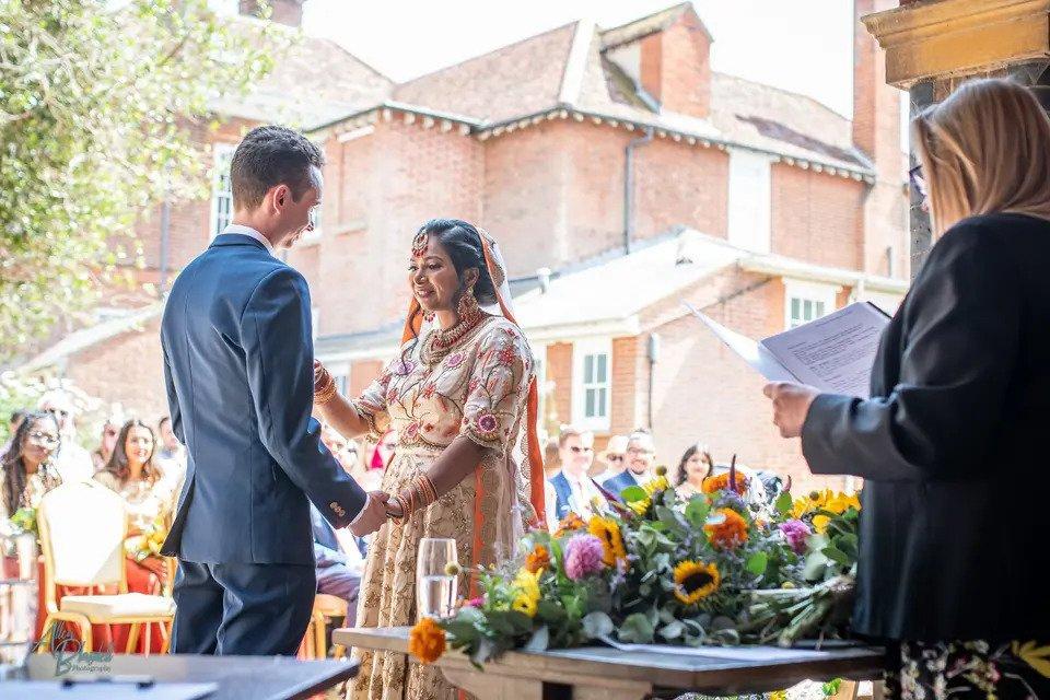 Bride and groom say their vows in an outdoor wedding ceremony in front of a floral arrangement. The groom is wearing a suit and the bride is dressed in traditional Asian wedding dress