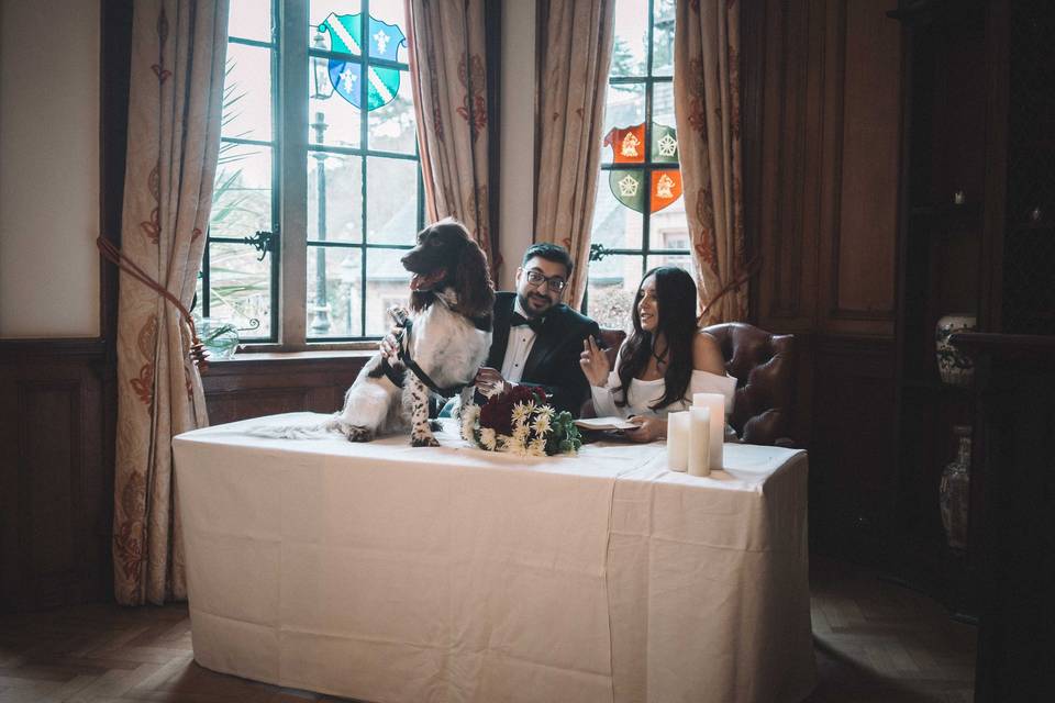 Couple sitting at a table at their wedding ceremony as they sign the register with Digby the Vid-dog-grapher sitting next to them
