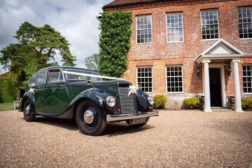 Green vintage wedding car with white bow parked on driveway of a red brick country house wedding venue