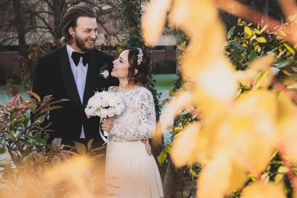 autumn leaves in the foreground as a bride and groom stand looking at one another in wedding attire