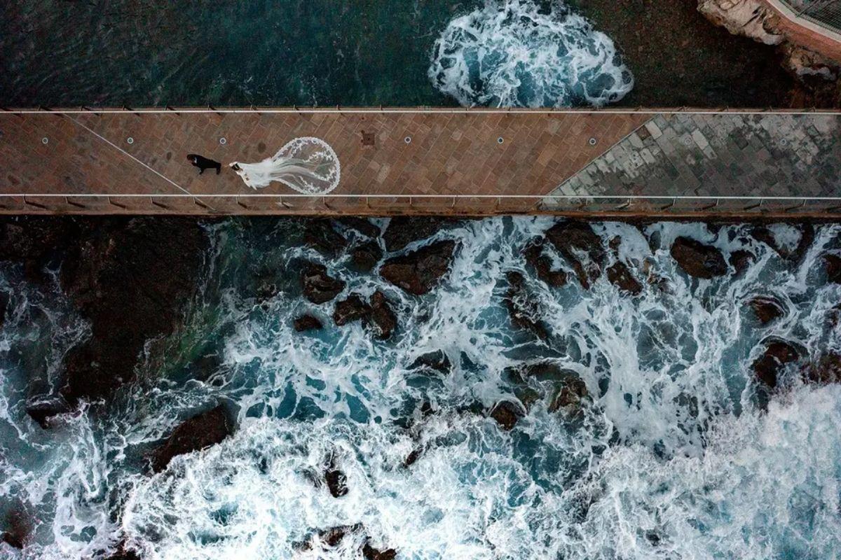 an aerial shot of a bride walking across a bride which sits above rough white waters