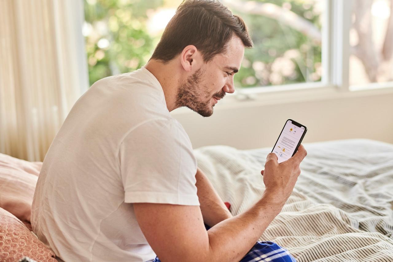 A man sat in his pyjamas on a bed reading a good morning text message on his phone.