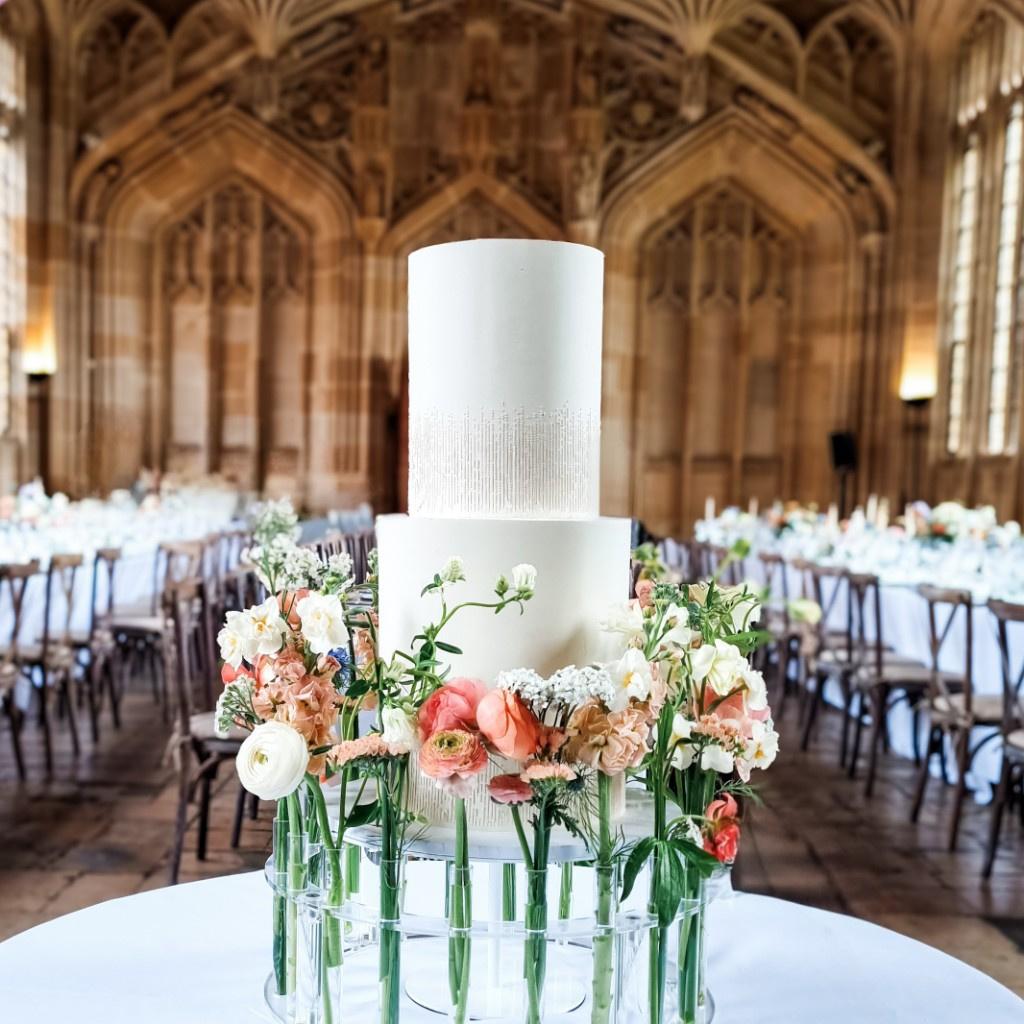 A two tier white simple wedding cake design on a clear cake stand decorated with fresh flowers.