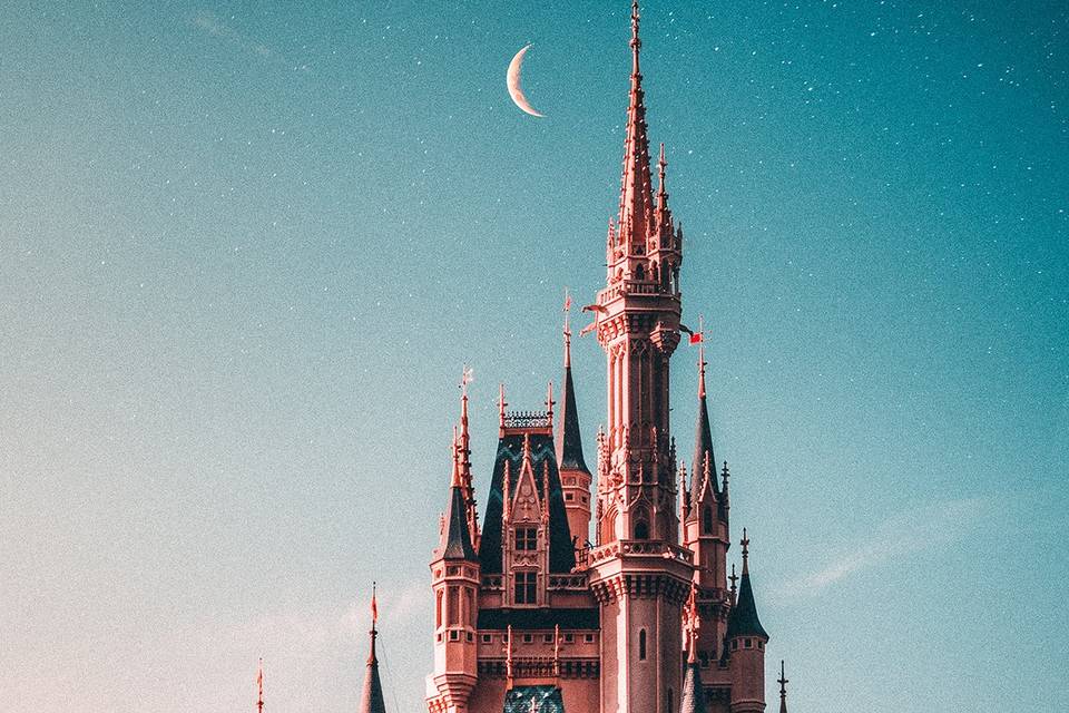 The turrets of a castle are silhouetted against a bright night sky, with a crescent moon to the left of the highest tower.