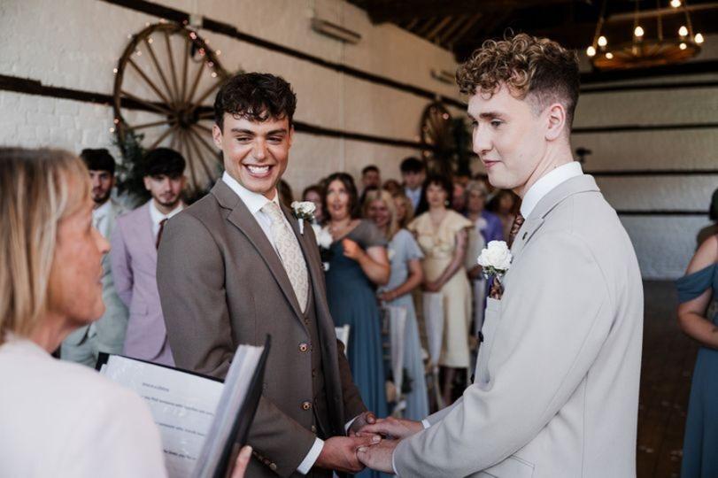 a couple wearing wedding suits holding hands at the altar as their celebrant reads from a binder