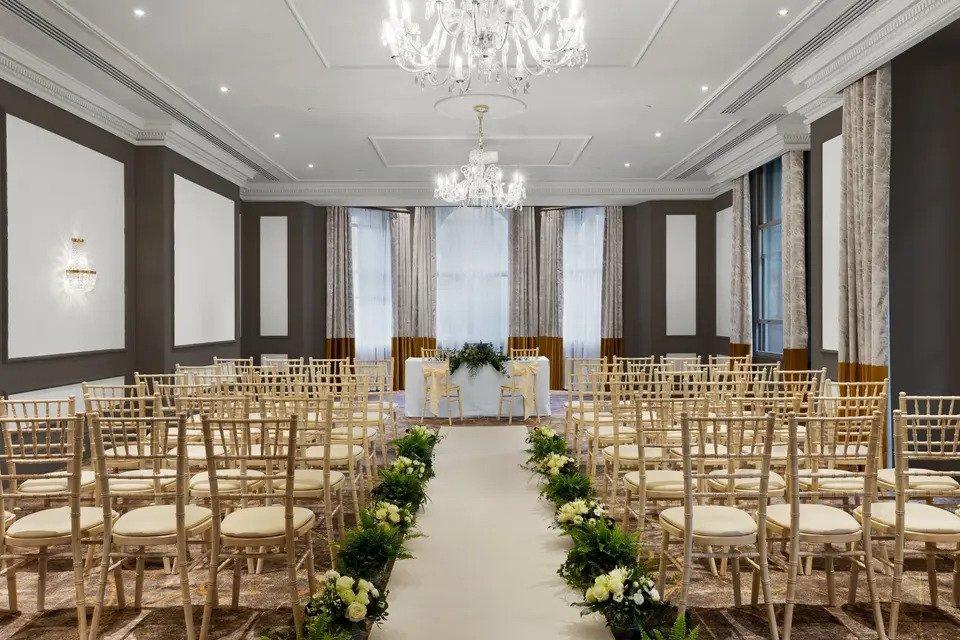 Hotel room set for wedding ceremony with Chiavari chairs, a flower-lined aisle with table, chandelier and drapes