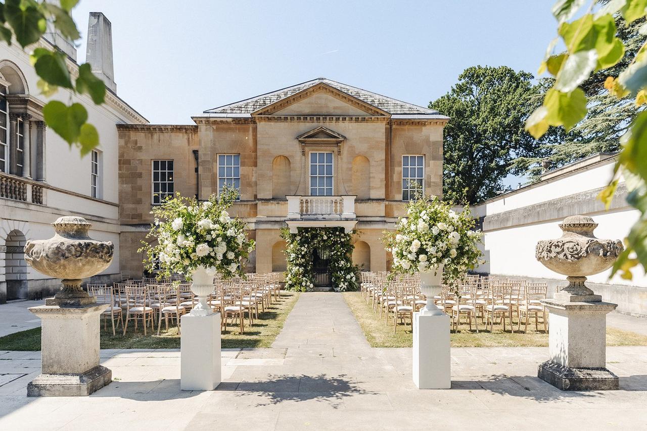 The exterior of a country house wedding venue courtyard set up for a wedding ceremony with plinth vases filled with white and green flowers and a fountain.