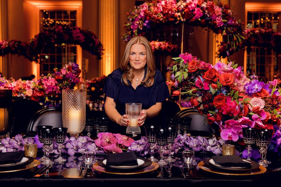 Sarah Haywood (white, blonde woman wearing a navy shirt and black trousers) seating among a display of pink and purple flowers in front of a laid table