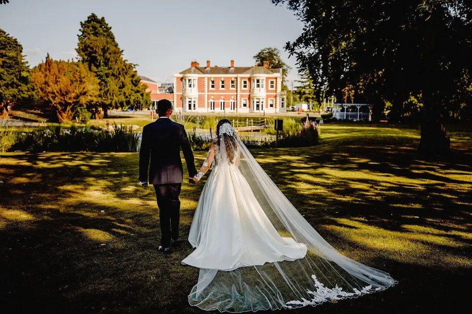 Bride and groom walk hand in hand towards a stately home wedding venue. The bride has a long skirt and veil