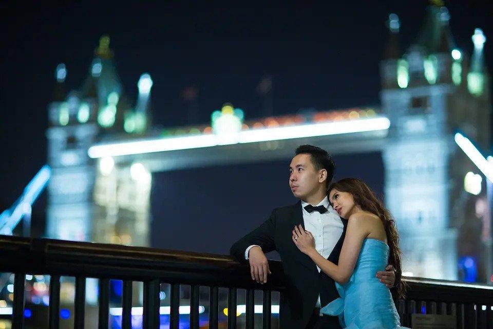 Bride and groom embrace and look into the distance in front of tower bridge at night