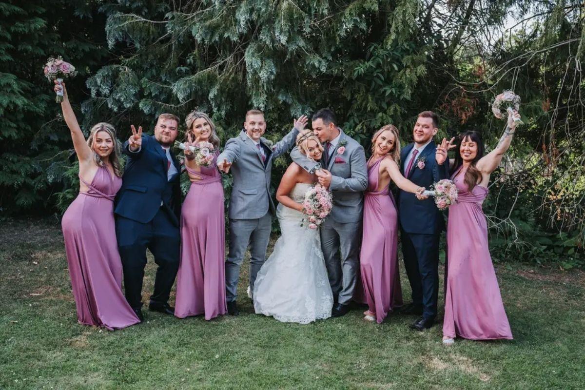 a bride and groom kiss in their venue's grounds as they pose with members of their wedding party in pink bridesmaid dresses and dark navy suits