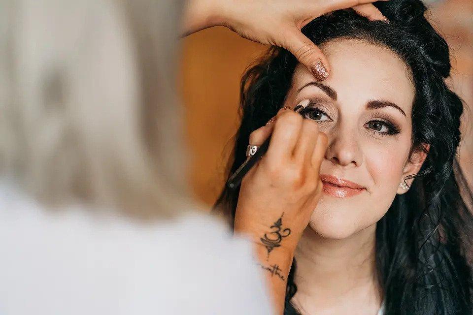 A smiling bride looks at an MUA applying eyeshadow in her crease