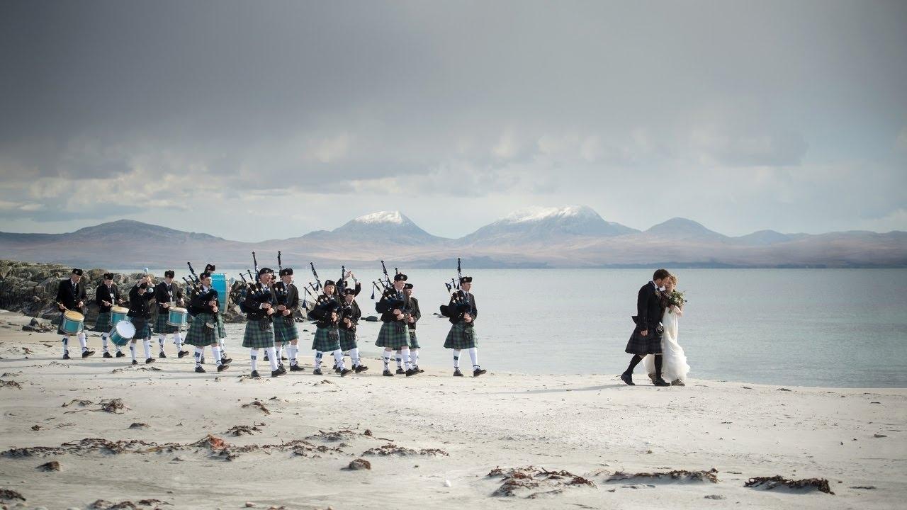 Couple in wedding attire walking along a beach in scotland followed by a series of scottish bagpipers and drummers
