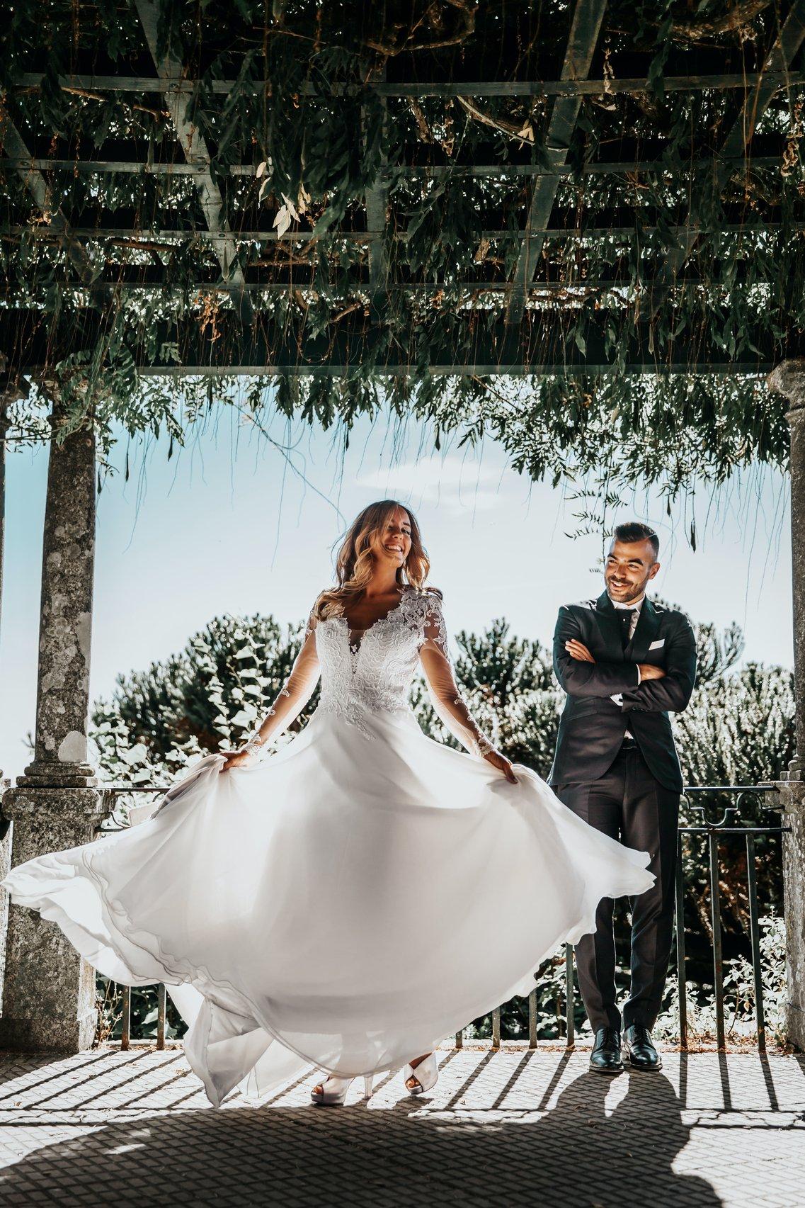 Laughing bride twirling her wedding dress while groom looks on laughing
