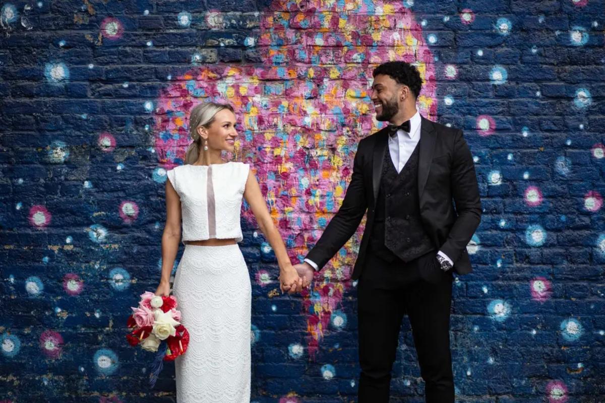  a couple hand in hand outside a small london wedding venue standing in front of a blue brick wall with a colourful heart painted on it