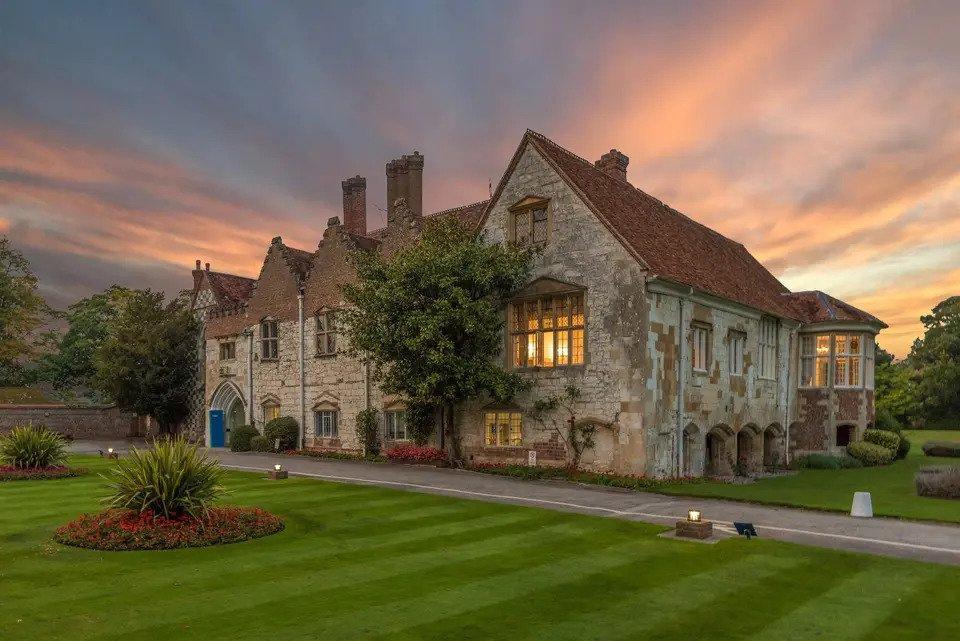 Grey stone building with red roof warmly lit from within photographed at dusk from across a manicured lawn