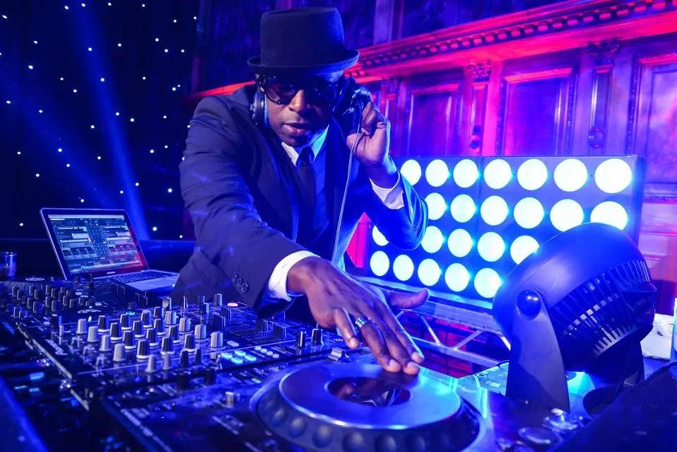A DJ wearing a suit, hat and sunglasses works the decks 