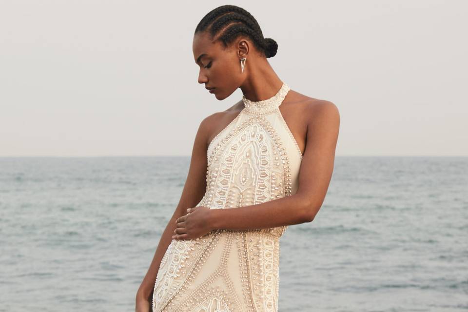 model looking to the side wearing a halterneck wedding dress in champagne colour covered in beading on a beach