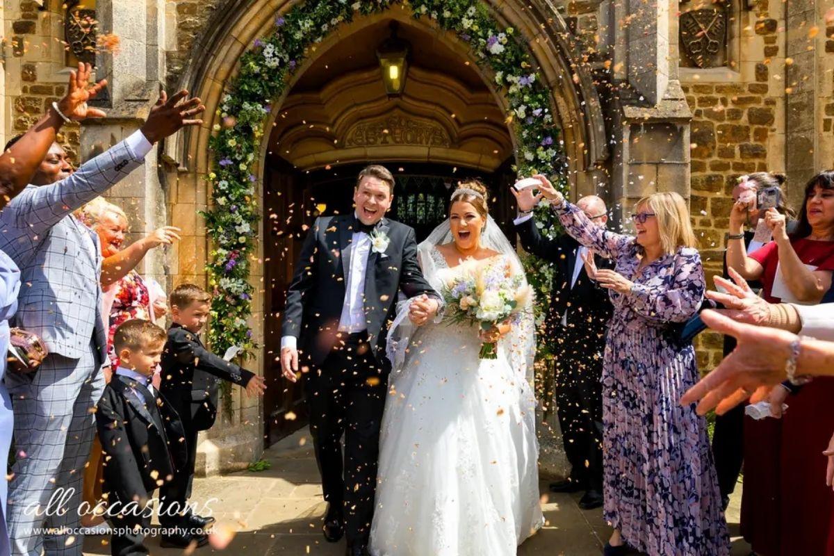 a bride and groom leaving their wedding ceremony with a floral arch behind them and wedding guests throwing confetti in front of them