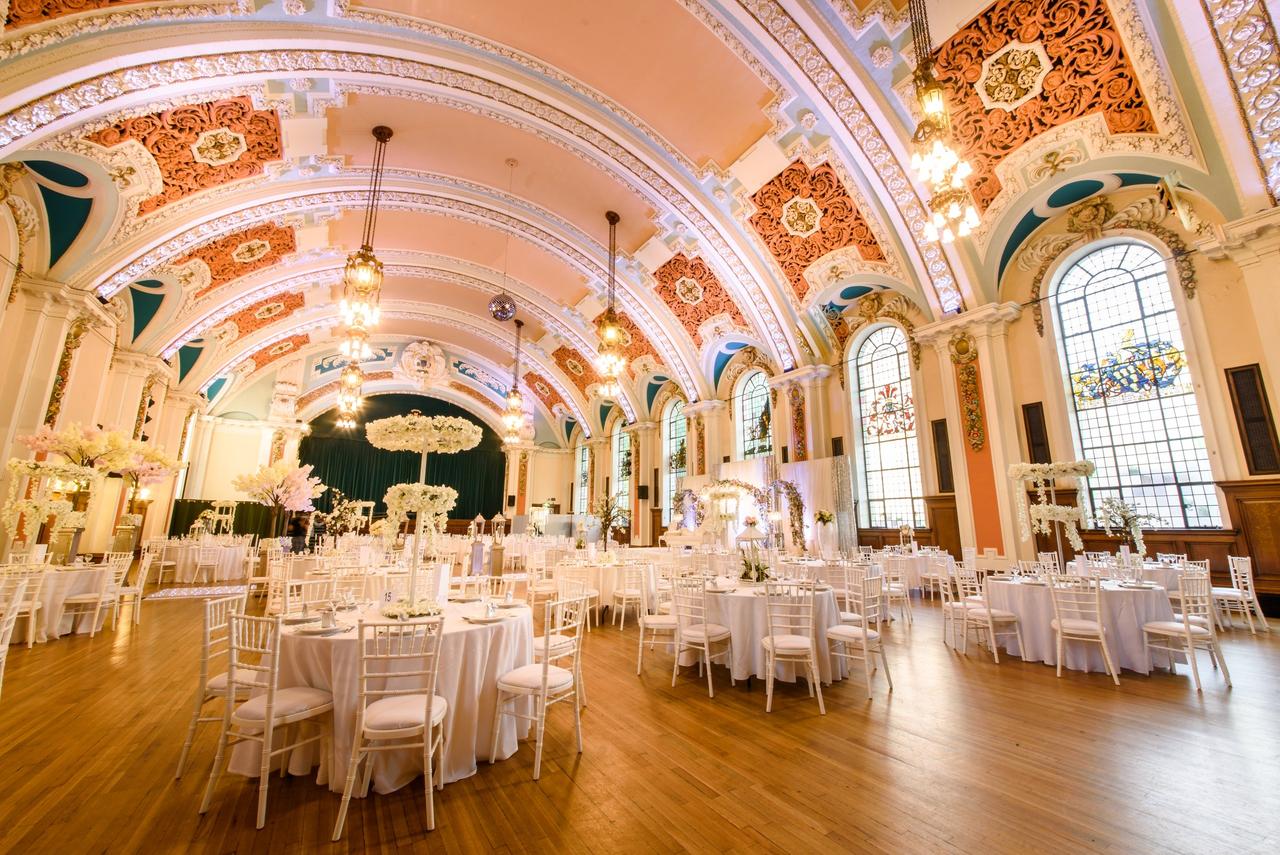 Interior of a ballroom set up for a wedding reception with high ceilings, large glass windows and round tables with tall opulent centrepieces.