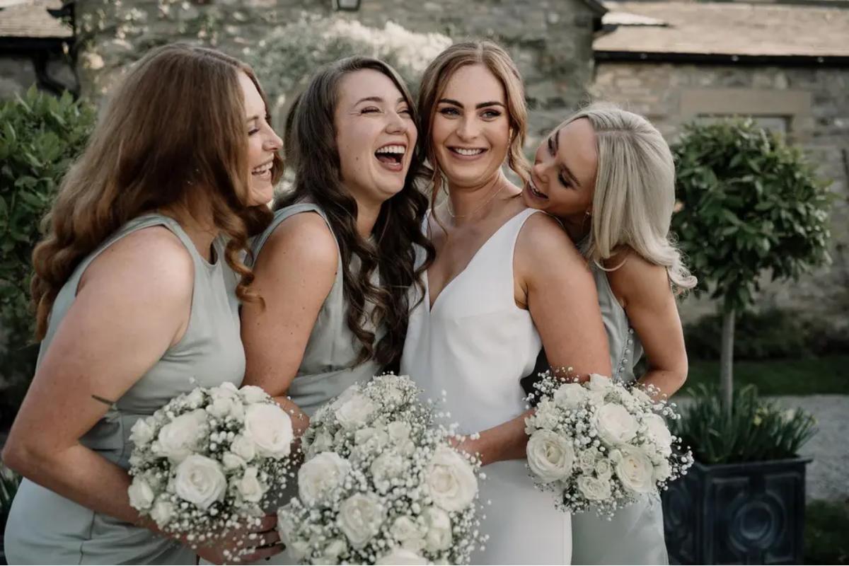 a bride with a low bun stood with her three bridesmaids wearing age green bridesmaid dresses smiling and holding bouquets - Alice Marie